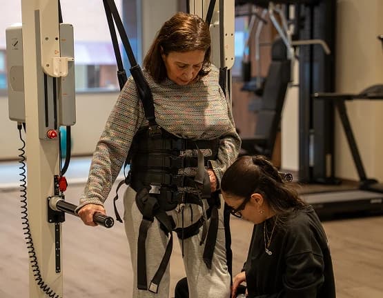 Imagen de una mujer caminando con un arnés de suspensión junto a una fisioterapeuta.
